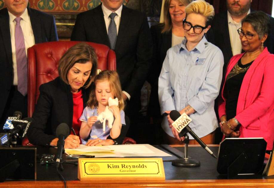 With Penelope Steele of Des Moines sitting in her lap, Iowa Gov. Kim Reynolds on Wednesday signs into law during a ceremony in the Iowa Capitol in Des Moines legislation that will require insurance companies to cover biomarker testing. (Erin Murphy/The Gazette)
