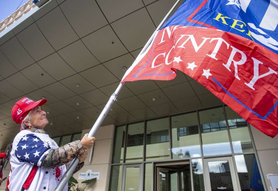 Julianna Balogh of Arkansas smiles as she holds out a large Trump 2024 flag for passing cars Saturday while waiting for his visit at the DoubleTree by Hilton Convention Complex in Cedar Rapids. Balogh said she has driven 60,000 miles this year to follow the former president around the country. (Savannah Blake/The Gazette)